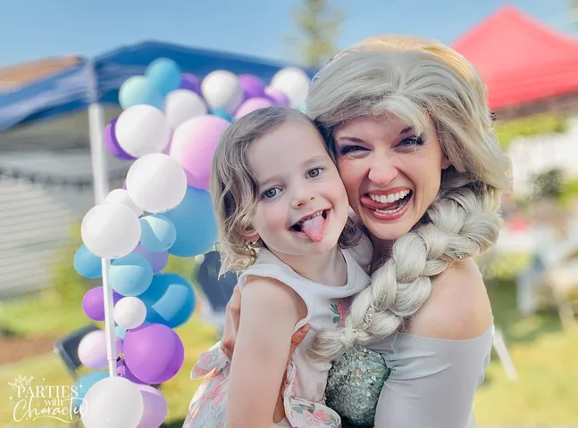 Elsa and a young girl sticking their tongues out beside a balloon garland outdoors