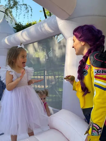 Rumi holding a microphone as a girl in a white tulle dress laughs inside a bounce house