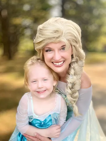 Elsa with her arms around a young girl in a sparkly blue dress among sunlit trees