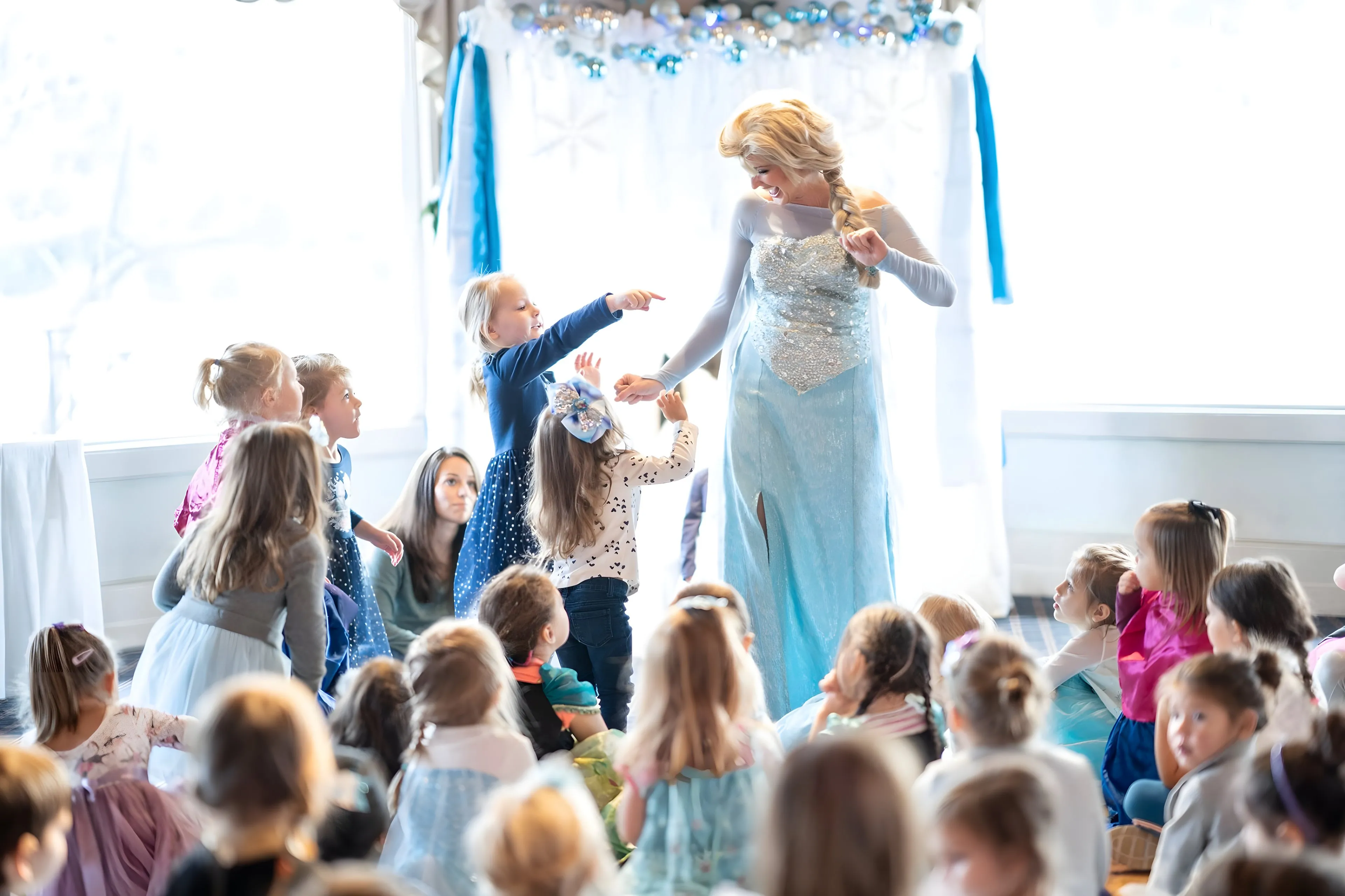 Jessica as Elsa in a bright room full of children, several reaching up toward her