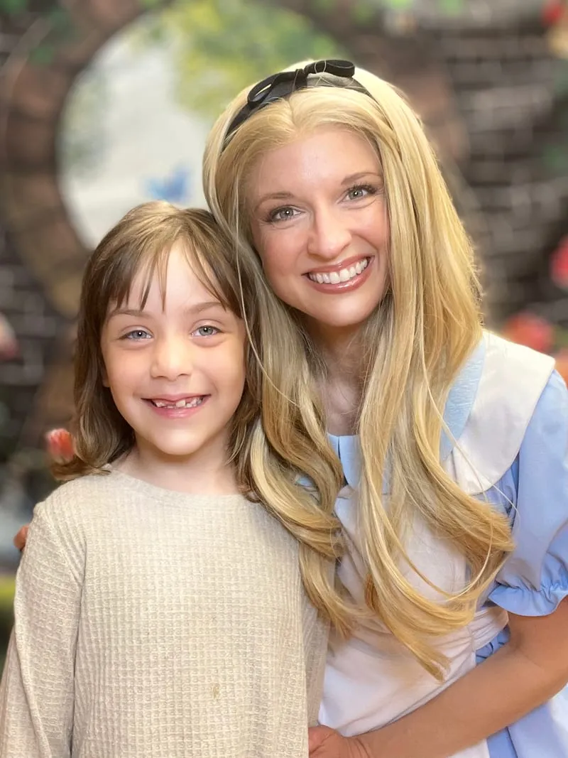 Alice with her arm around a smiling young girl in a cream sweater in front of a painted Wonderland backdrop at a library event