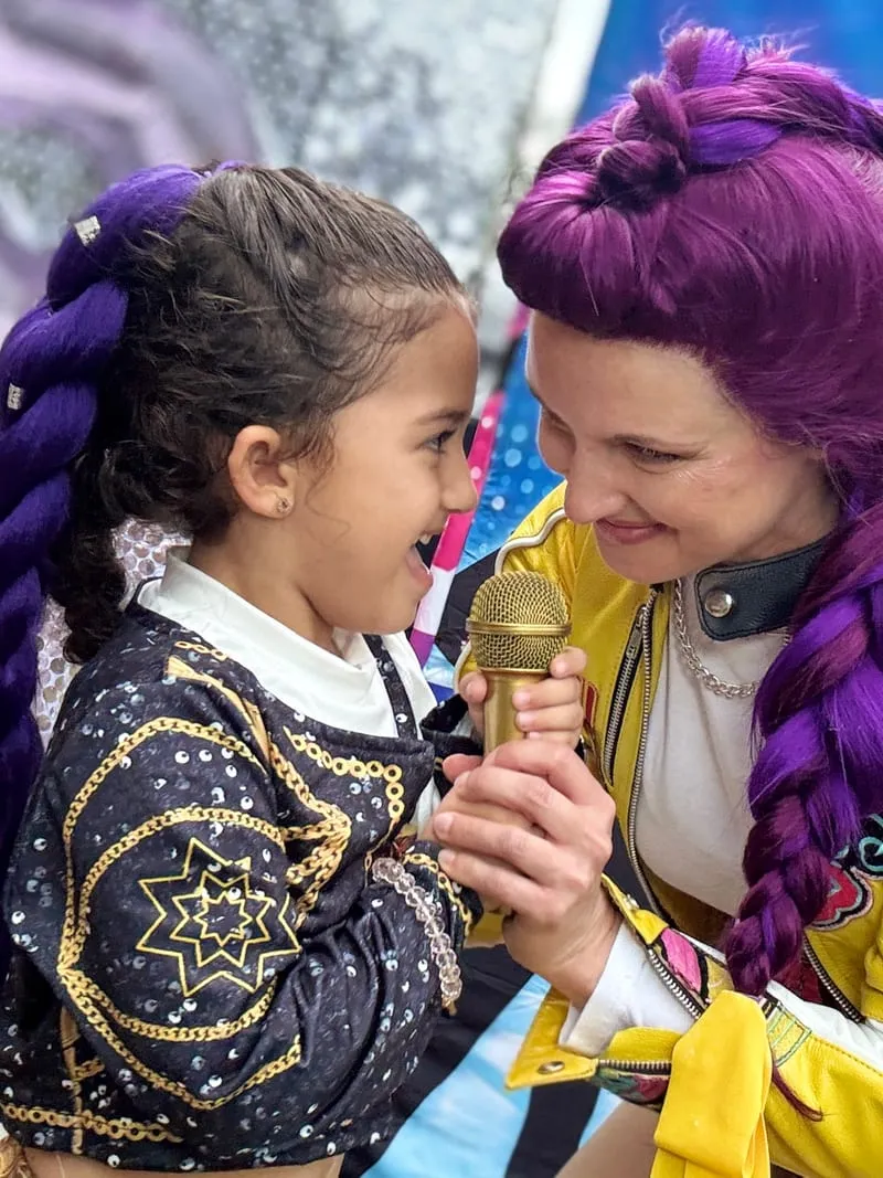 Rumi singing into a gold microphone face to face with a young girl wearing purple braids