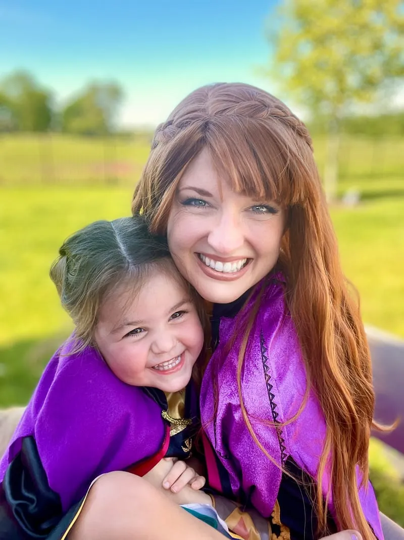 Anna cuddling a laughing young girl in a matching purple cape on a sunny lawn