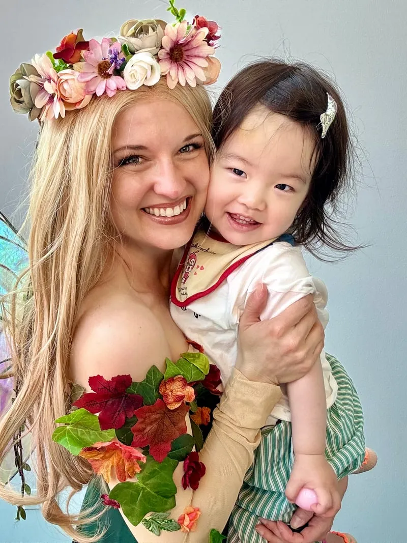 Woodland Fairy in a flower crown holding a smiling toddler girl in a white tee against a plain grey wall