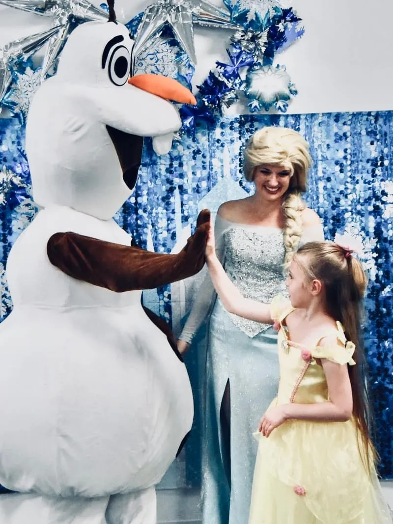 Olaf high-fiving a young girl in a yellow Belle dress while Elsa looks on in front of a snowflake backdrop