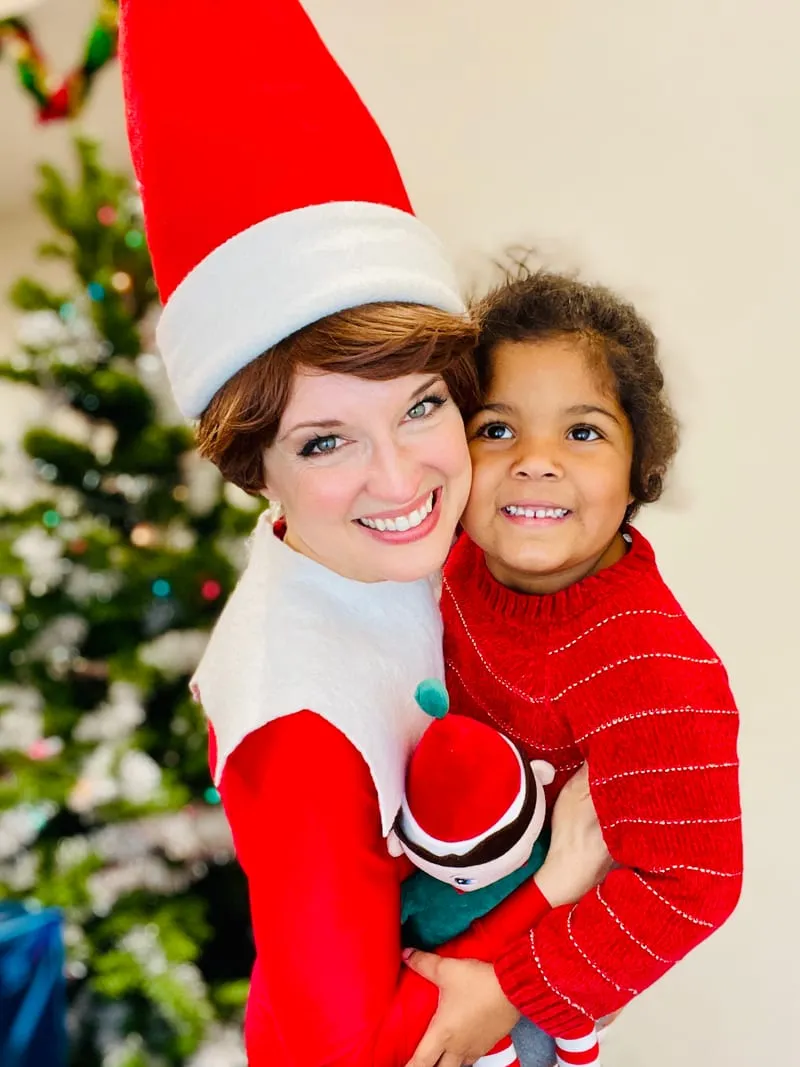 Elf on the Shelf cradling a young girl holding a candy cane by a Christmas tree