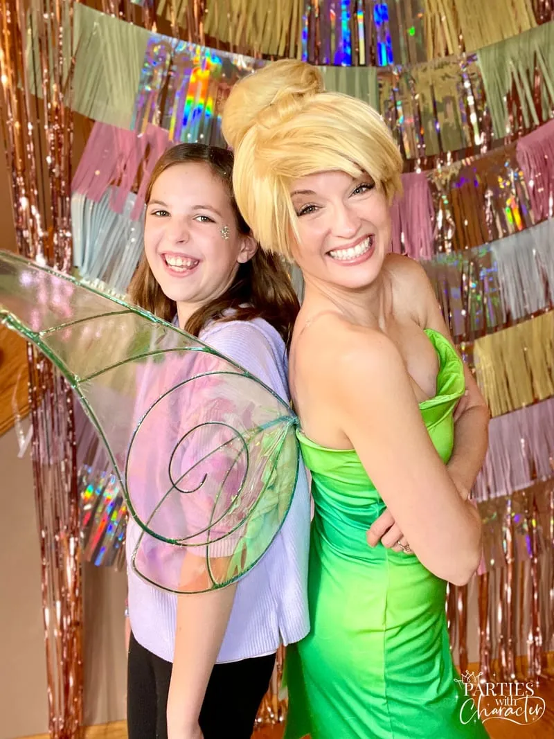 Tinker Bell standing back to back with a laughing girl in a lavender top in front of a pastel fringe backdrop